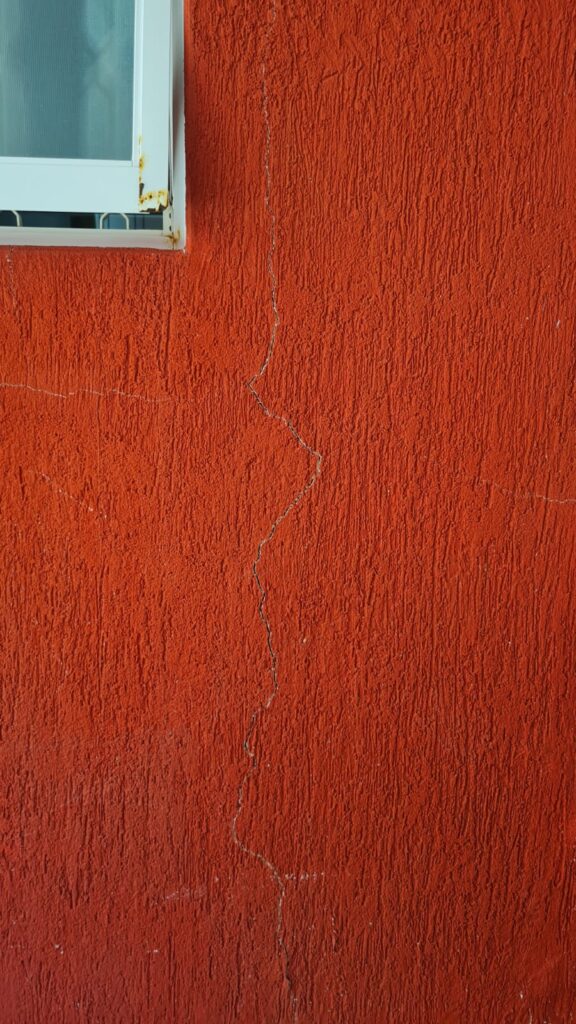 Fissura vertical em fachada com revestimento texturizado vermelho, próxima à esquadria, identificada durante vistoria técnica.