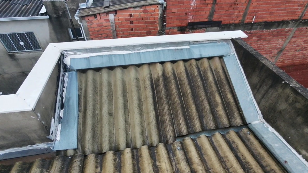 Vista aérea capturada por drone de uma cobertura com telhas onduladas de fibrocimento desgastadas e escurecidas pela ação do tempo, destacando o sistema de rufos metálicos e mantas aluminizadas de vedação nas bordas para prevenção de infiltrações.
