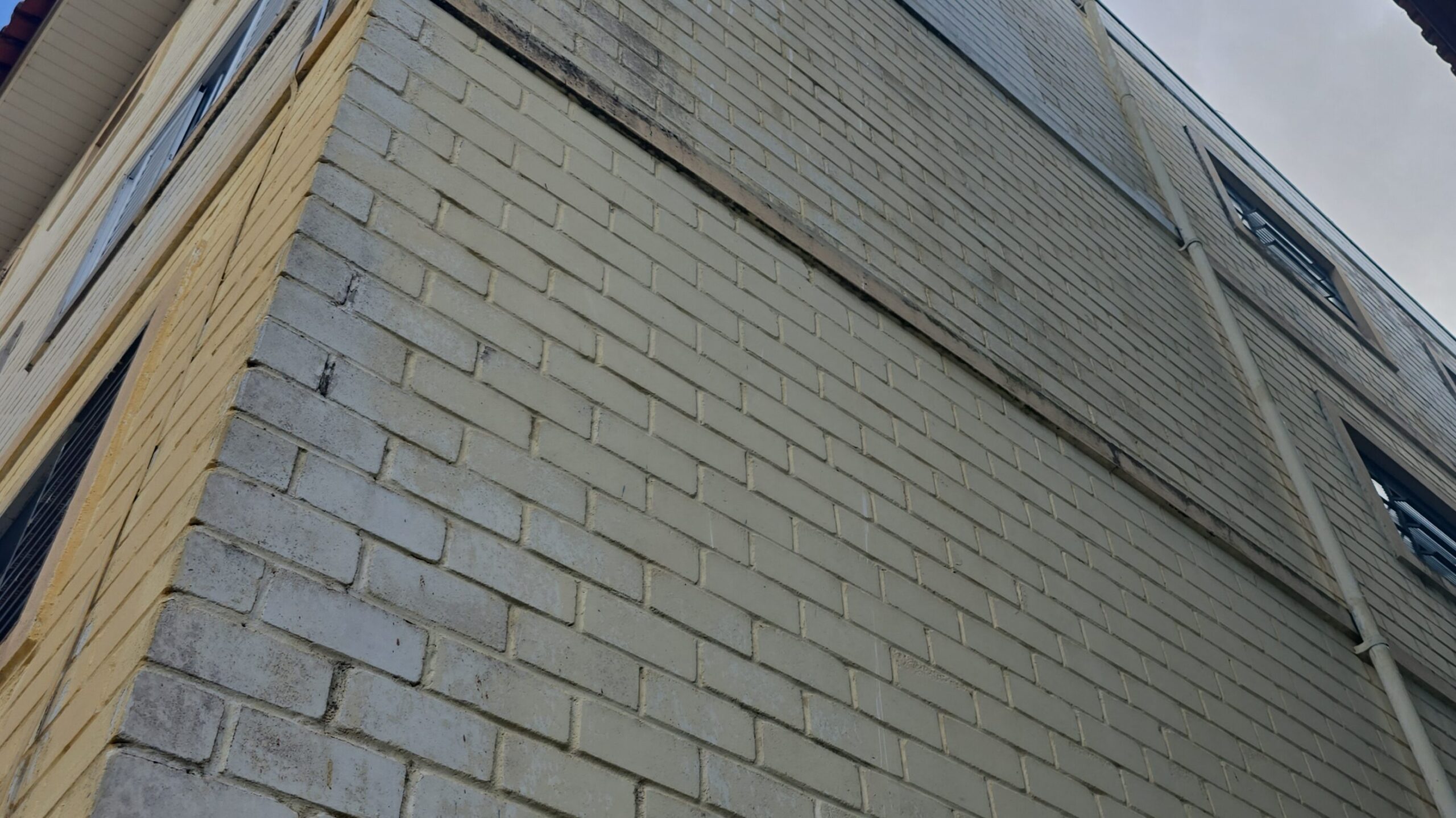 Vista em perspectiva inferior de uma fachada externa de edifício residencial revestida com blocos ou tijolos cerâmicos pintados de claro, apresentando janelas escuras e tubulação aparente de escoamento vertical sob céu com nuvens